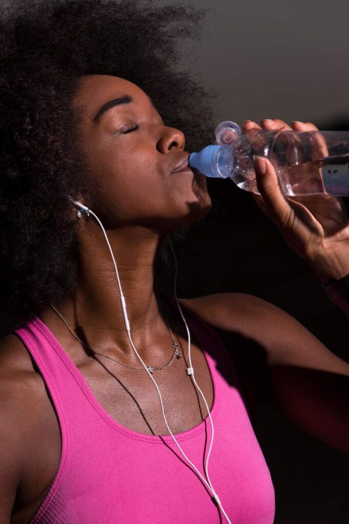 Young beautiful african american woman with headphones drinking water after jogging in nature beautiful summer night