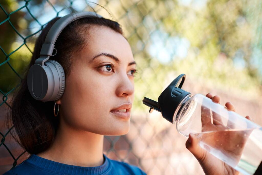 Shot of a sporty young woman drinking water while standing against a fence outdoors.