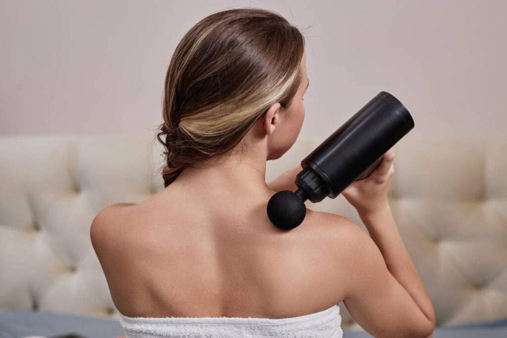 Back view of young woman using massager for shoulders at home