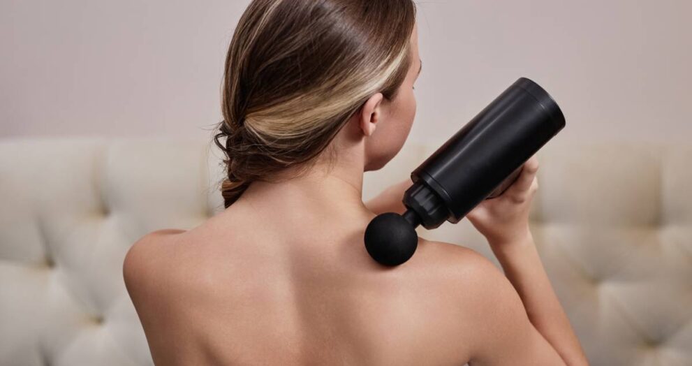 Back view of young woman using massager for shoulders at home